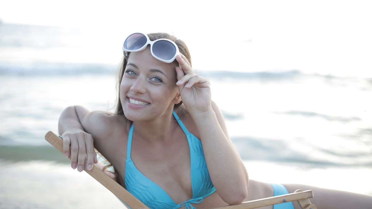 A woman in a blue swimsuit enjoys a sunny day at the beach, reclining on a folding chair with sunglasses.